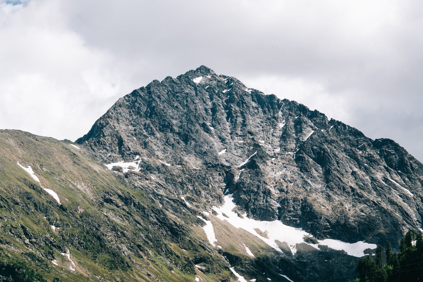 A rugged, dark grey mountain peak with patches of white snow in its crevices, set against a cloudy, overcast sky.