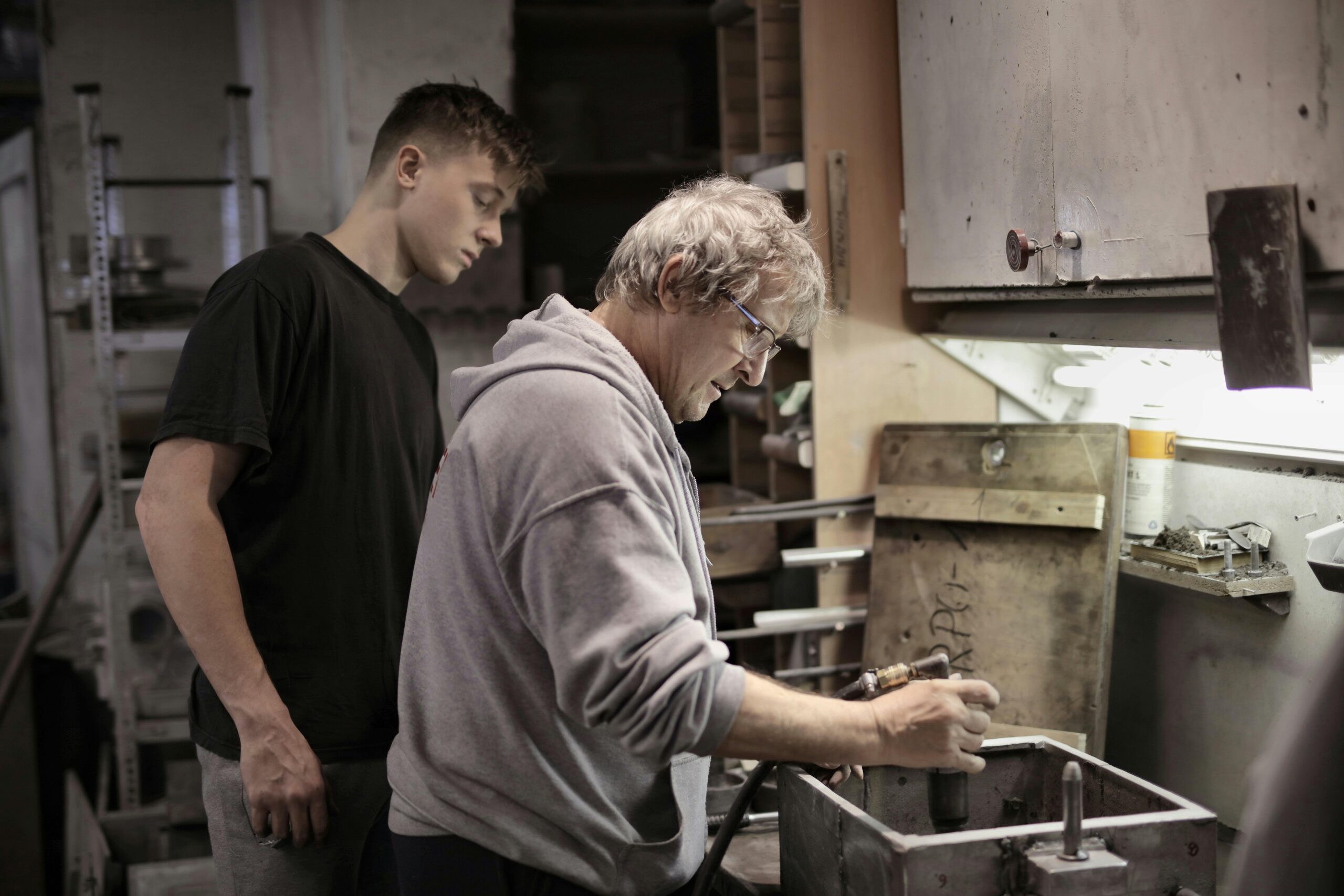 Older craftsperson demonstrating hands-on workshop work to a younger learner at a workbench, suggesting intergenerational skills transfer and apprenticeship training.