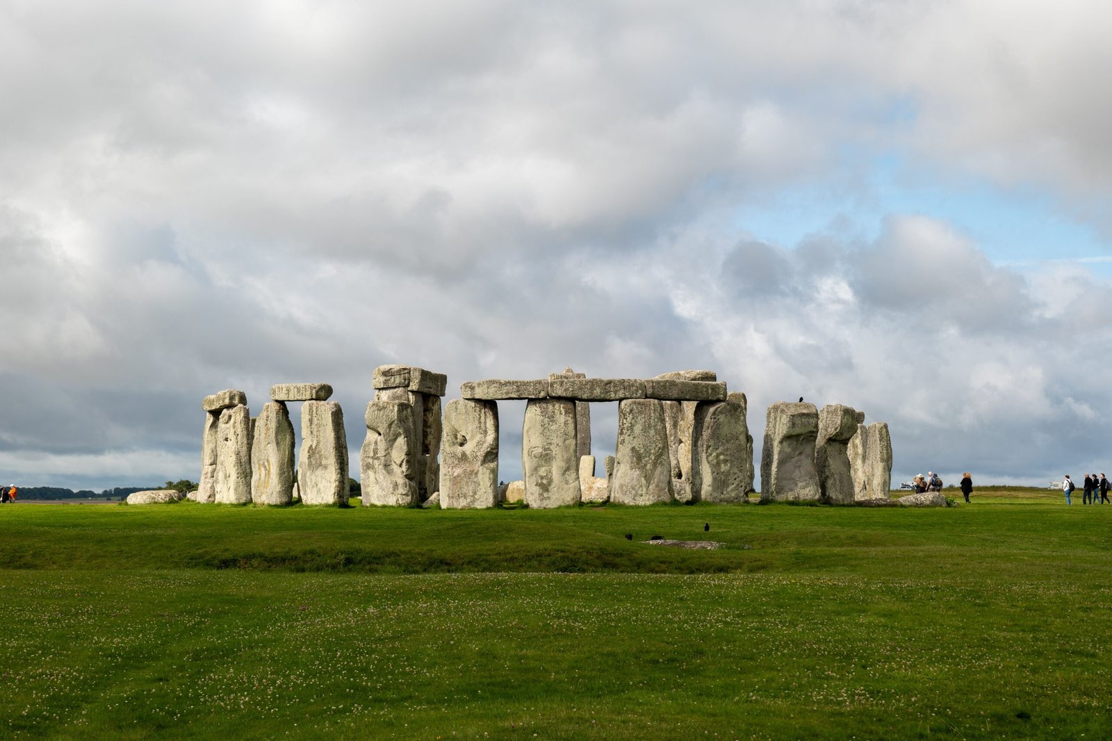 Stonehenge standing on a broad green lawn beneath dramatic grey clouds, with patches of blue sky and a few visitors visible in the distance.
