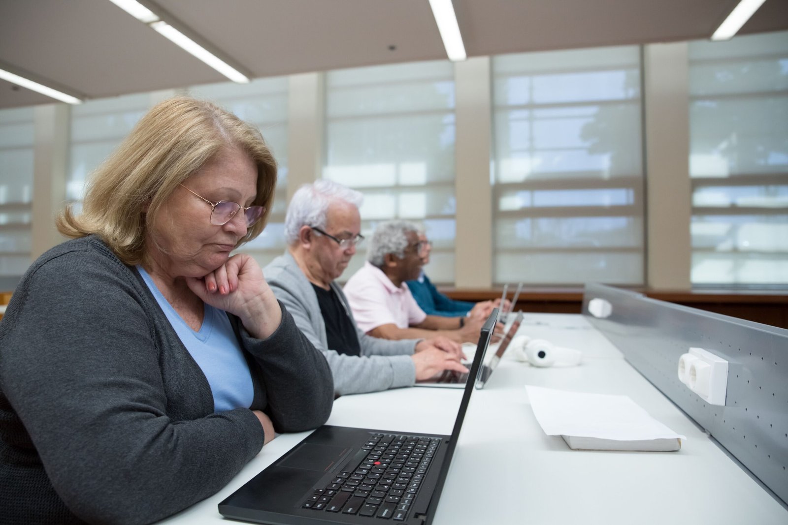 Four older adults are seated in a row along a white desk in a bright, modern room, all using laptops. In the foreground, a woman with blonde hair and glasses rests her chin on her hand, looking thoughtfully at her screen.